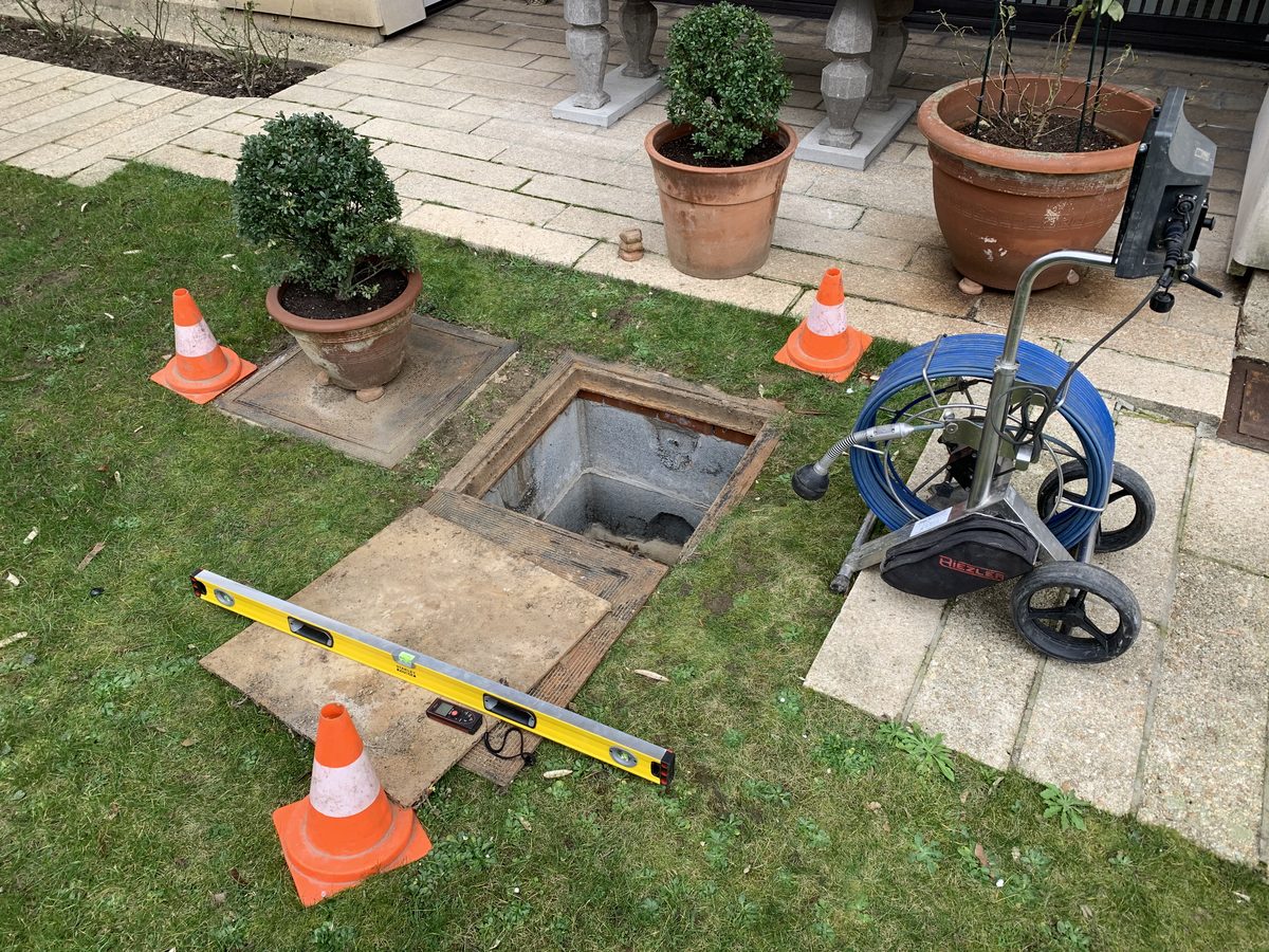 Chambre de visite ouverte dans un jardin, enrouleur de caméra d'inspection et niveau posés à côté, zone balisée par des cônes.